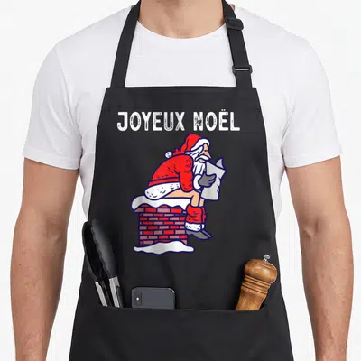Tablier de cuisine noir avec illustration colorée d'un Père Noël assis sur une cheminée en train de lire un papier.