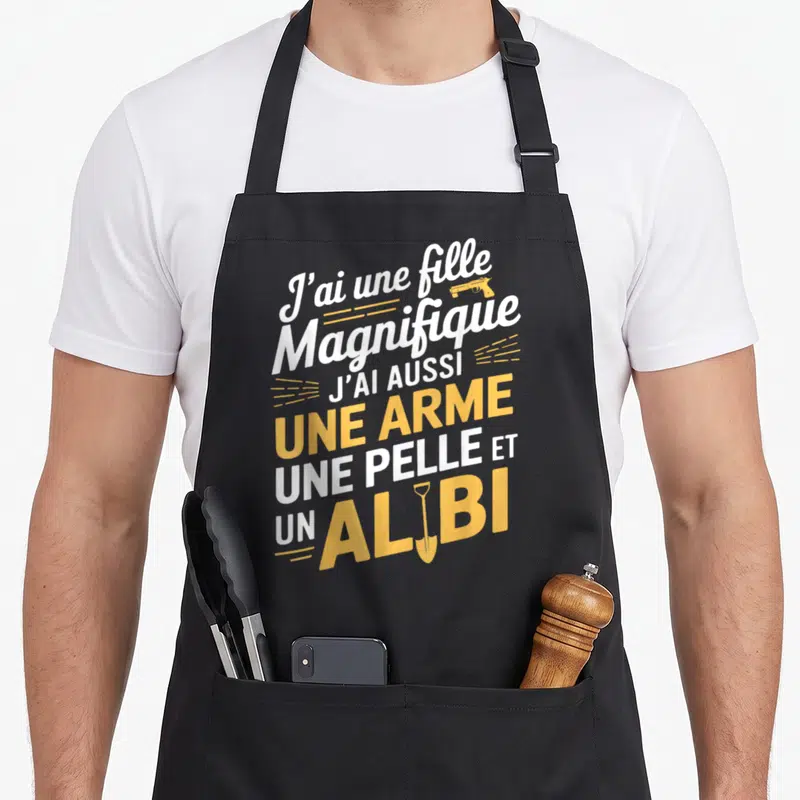 Tablier de cuisine noir avec inscription blanche et jaune J'ai une fille magnifique une arme une pelle et un alibi illustré.