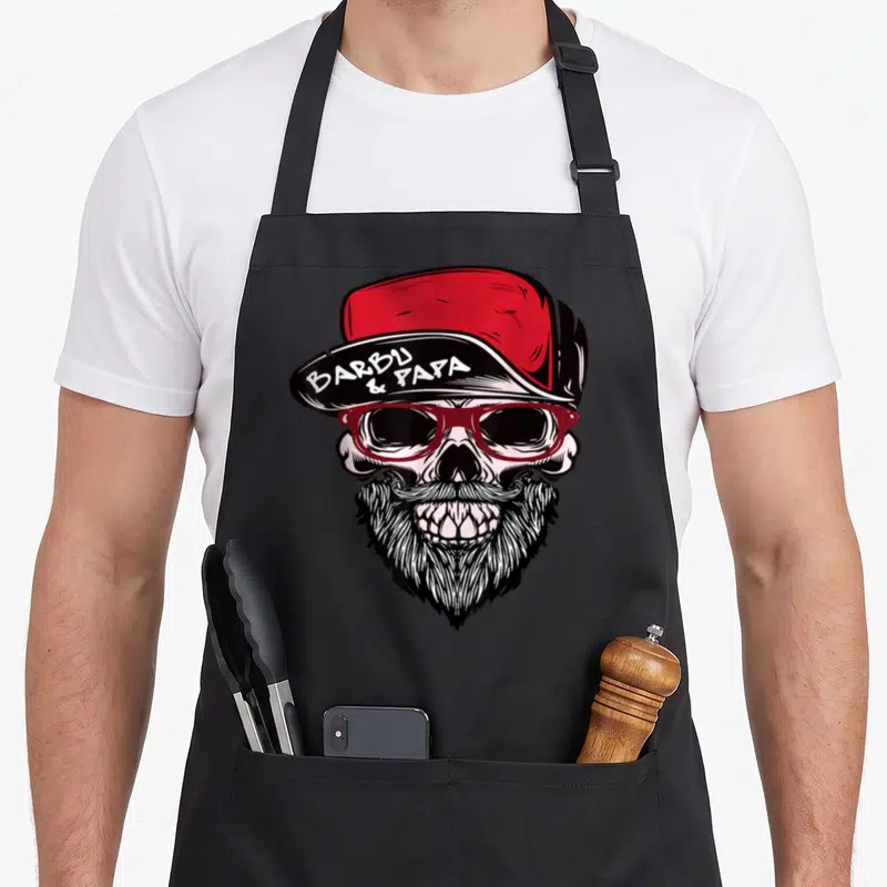 Tablier de cuisine noir avec un crâne barbu portant des lunettes rouges et une casquette inscrite Barbu & Papa.