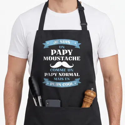 Tablier de cuisine noir avec inscription Je suis un papy moustache et illustration de moustache blanche sur fond bleu clair.