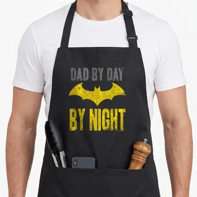 Tablier de cuisine noir avec emblème de chauve-souris jaune et texte Dad By Day By Night imprimé en lettres capitales.