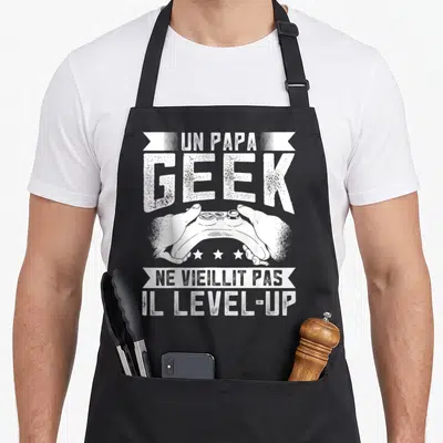 Tablier de cuisine noir avec un Papa Geek, une manette de jeu vidéo et trois étoiles blanches imprimés au centre.