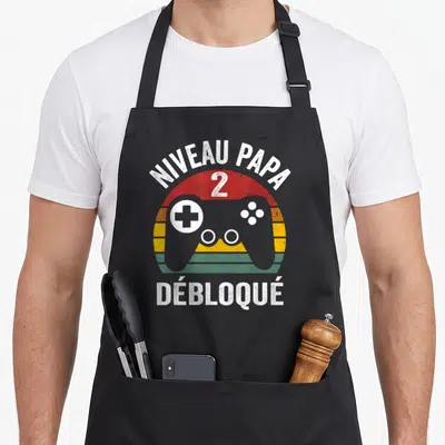 Tablier de cuisine noir avec illustration manette de jeu vidéo et texte Niveau Papa 2 Débloqué en style rétro.