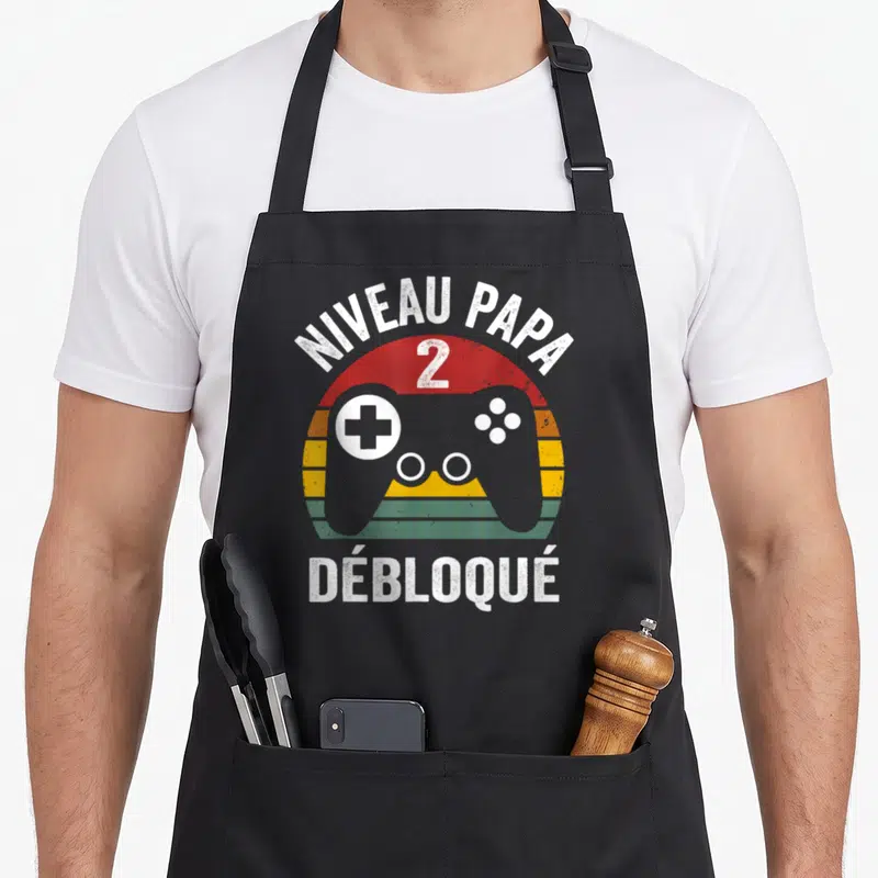Tablier de cuisine noir avec illustration manette de jeu vidéo et texte Niveau Papa 2 Débloqué en style rétro.