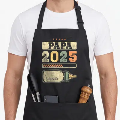 Tablier de cuisine noir avec inscription Papa 2025 et illustration de biberon vintage coloré.