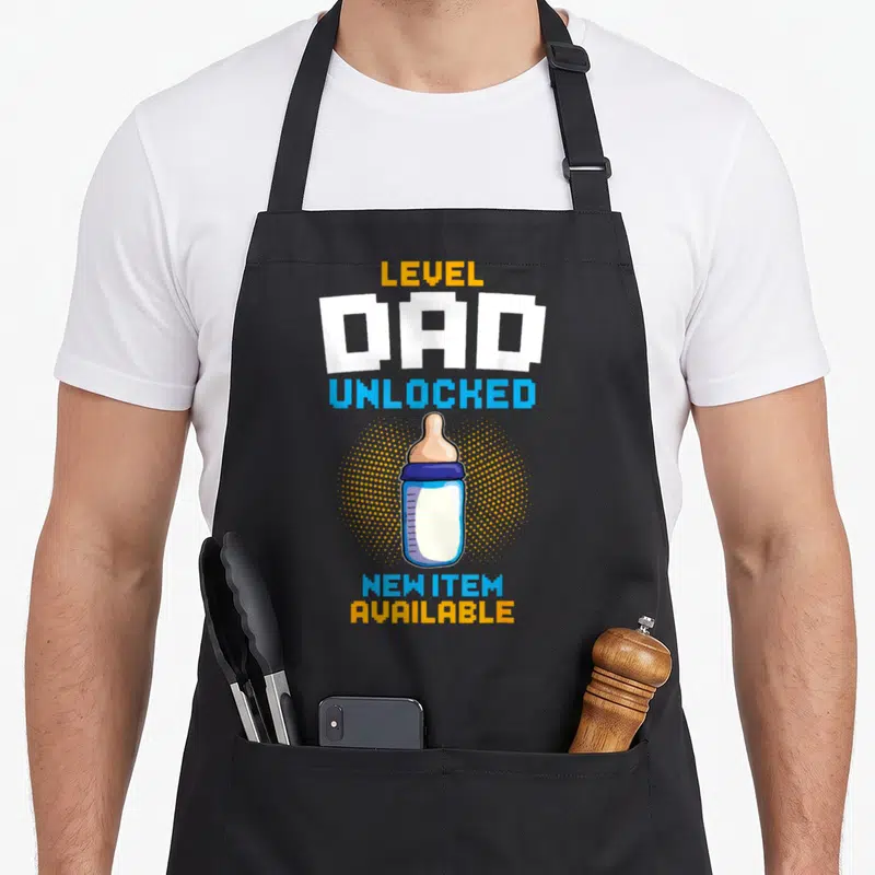 Tablier de cuisine noir avec une illustration de biberon et le texte Level Dad Unlocked New Item Available en couleurs vives.