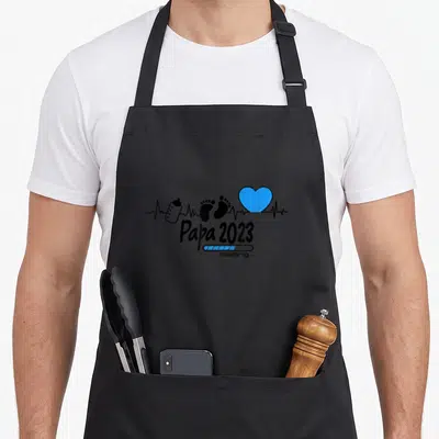 Tablier de cuisine noir avec inscription Papa 2023, biberon, traces de pas et cœur bleu sur fond de ligne cardiaque.