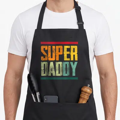 Tablier de cuisine noir avec l'inscription Super Daddy en typographie colorée et effet vieilli, encadrée par deux bandes horizontales.