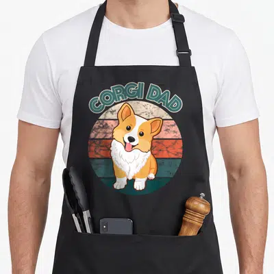 Tablier de cuisine noir avec un corgi cartoon souriant et l'inscription Corgi Dad en arc de cercle au-dessus.