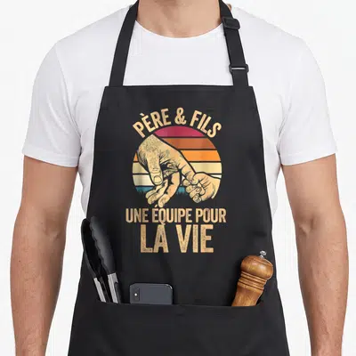 Tablier de cuisine noir avec une illustration vintage représentant une main d'adulte tenant celle d'un bébé devant un soleil aux couleurs chaudes.