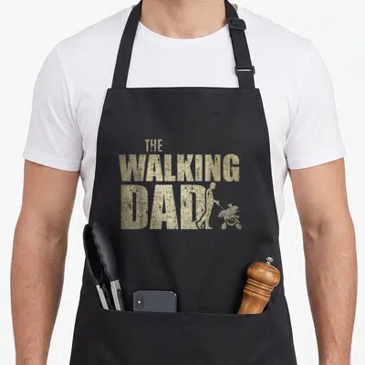 Tablier de cuisine noir avec le texte The Walking Dad et une silhouette poussant une poussette en doré texturé.