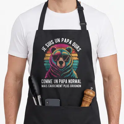 Tablier de cuisine noir avec illustration colorée d'un ours portant des lunettes et un texte humoristique en blanc.