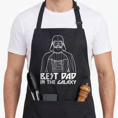Tablier de cuisine noir avec une illustration blanche de Darth Vader et le texte Best Dad In The Galaxy en bas du motif.