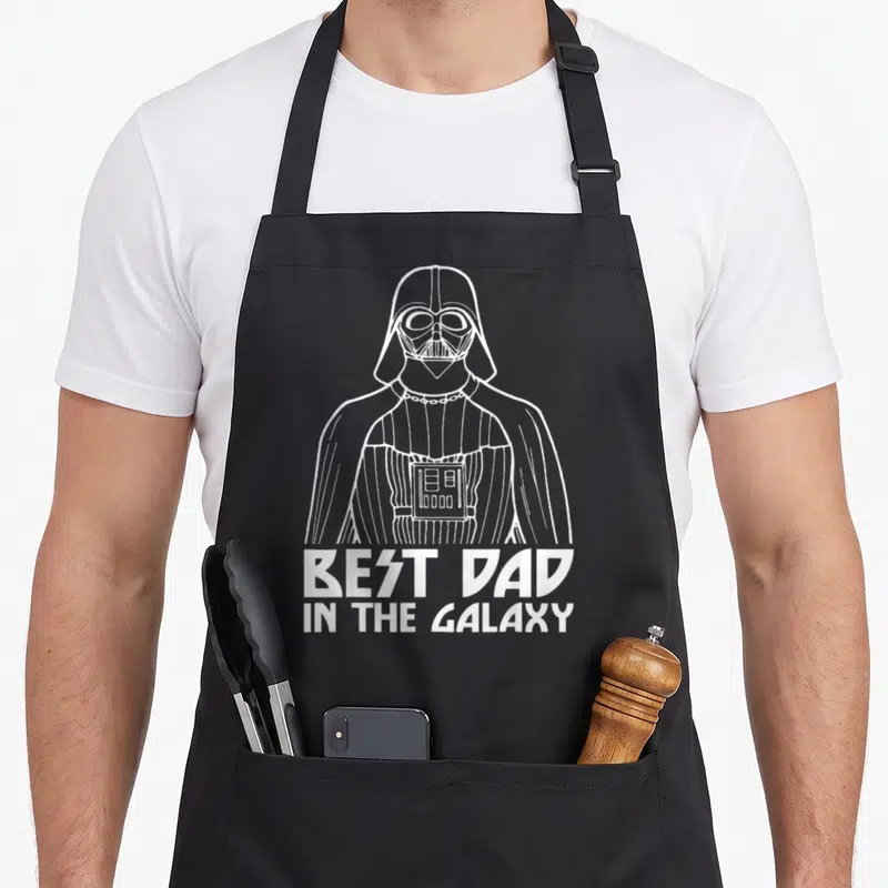 Tablier de cuisine noir avec une illustration blanche de Darth Vader et le texte Best Dad In The Galaxy en bas du motif.