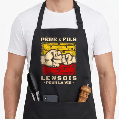 Tablier de cuisine noir avec l'inscription Père et Fils Lensois et deux poings qui se touchent sur un fond jaune et rouge.