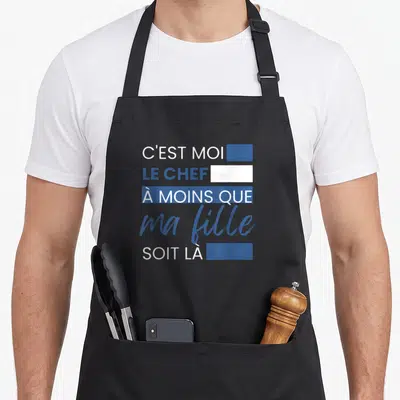 Tablier de cuisine noir avec inscription C'est moi le chef à moins que ma fille soit là en typographie blanche et bleue.