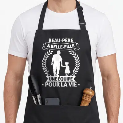 Tablier de cuisine noir avec une silhouette blanche d'un homme et d'une enfant tenant la main, entourés de couronnes végétales stylisées.