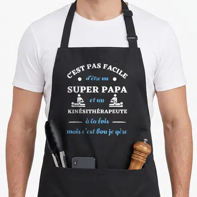 Tablier de cuisine noir avec texte blanc et bleu C'est pas facile d'être un super papa et un kinésithérapeute à la fois mais c'est bon je gère.