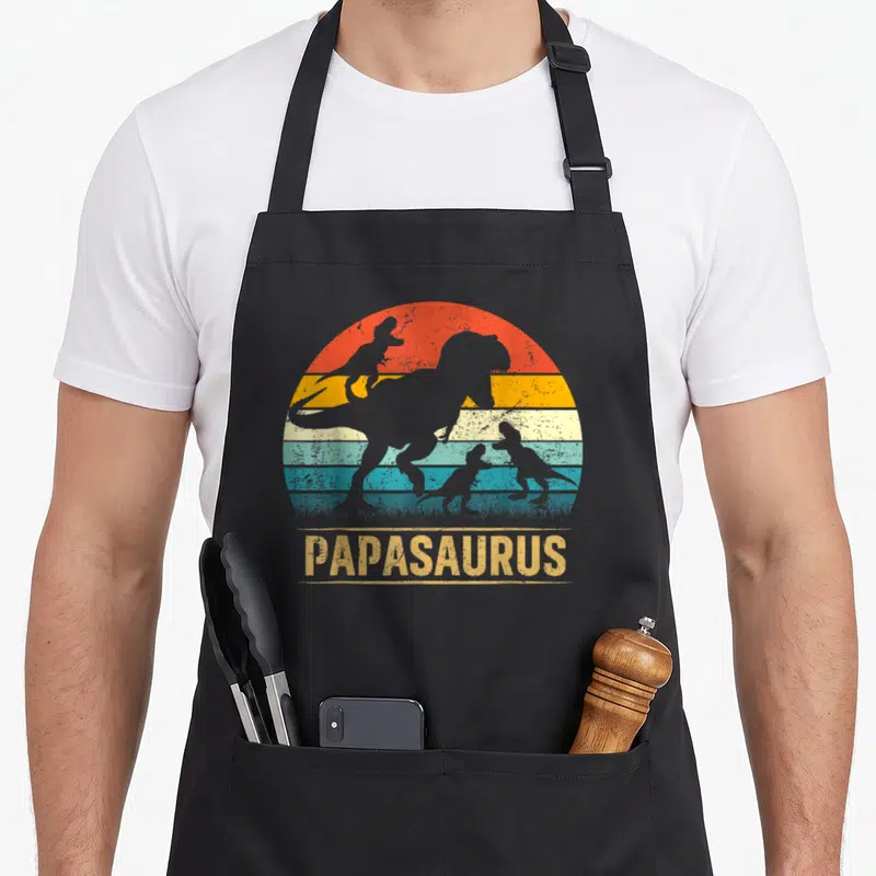 Tablier de cuisine noir avec illustration vintage Papasaurus et silhouettes de dinosaures.