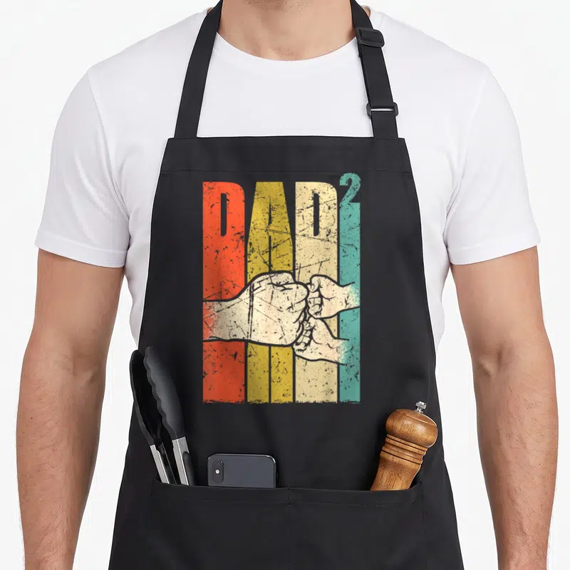 Tablier de cuisine noir avec inscription Dad² colorée et illustration de deux poings qui se tapent amicalement.