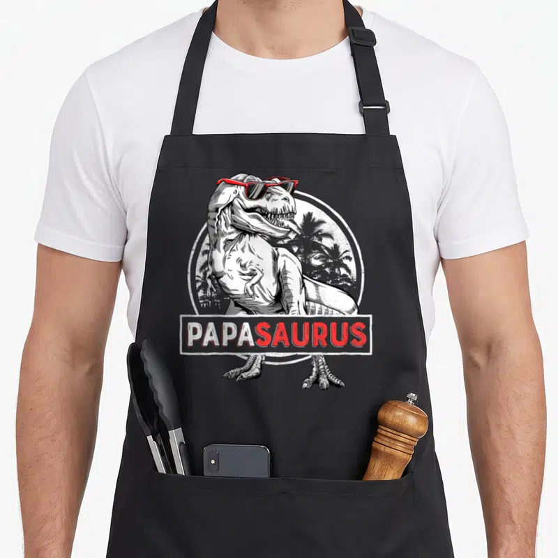 Tablier de cuisine noir avec illustration d'un T-Rex portant des lunettes de soleil rouges et le mot Papasaurus.
