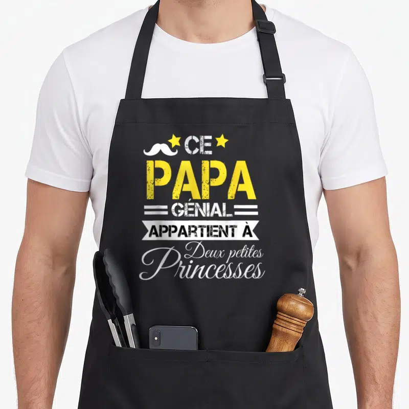 Tablier de cuisine noir avec une moustache blanche, deux étoiles jaunes et le texte Ce Papa Génial Appartient à Deux Petites Princesses.