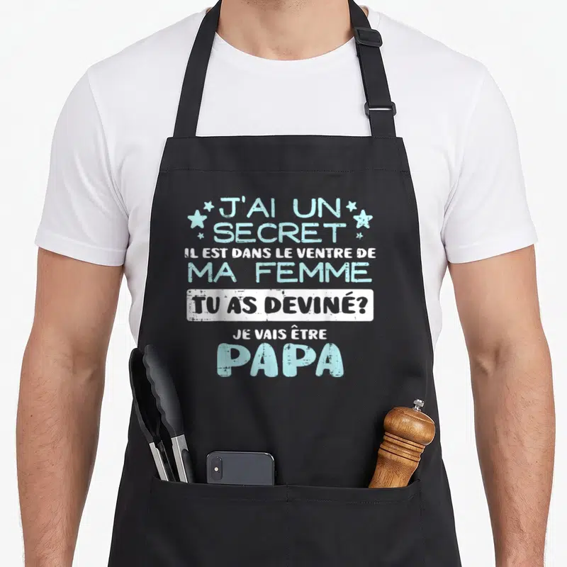 Tablier de cuisine noir avec inscription bleue et blanche annonçant la naissance d'un enfant et la future paternité.