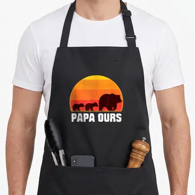 Tablier de cuisine noir avec une illustration rétro montrant un grand ours suivi de deux petits oursons devant un soleil orange.