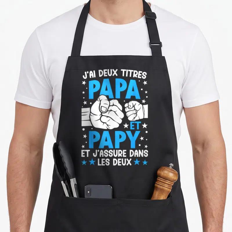 Tablier de cuisine noir avec poings serrés et texte J'ai deux titres Papa et Papy entouré d'étoiles bleues et blanches.