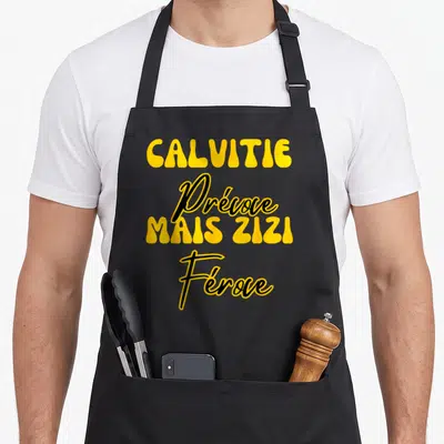 Tablier de cuisine noir avec le texte jaune Calvitie Prévoe Mais Zizi Féroce imprimé au centre du torse.