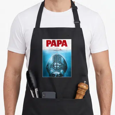 Tablier de cuisine noir avec une illustration parodique montrant un crâne sous l'eau et le mot PAPA en lettres rouges au sommet.
