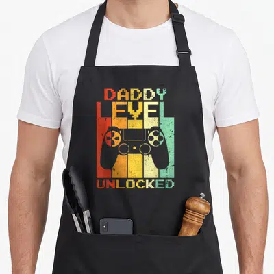 Tablier de cuisine noir avec inscription Daddy Level Unlocked et silhouette de manette de jeu vidéo sur fond de bandes colorées pixelisées