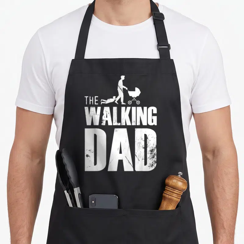 Tablier de cuisine noir avec silhouette père poussette enfant traîné et texte The Walking Dad en blanc.
