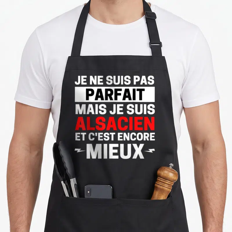 Tablier de cuisine noir avec le texte Je ne suis pas parfait mais je suis Alsacien et c'est encore mieux imprimé en blanc et rouge.