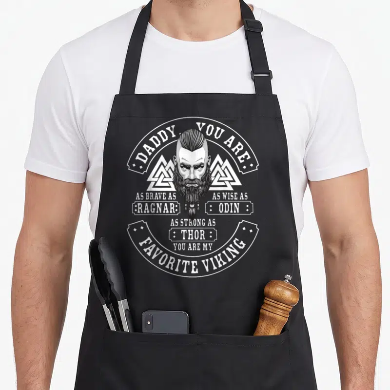 Tablier de cuisine noir avec illustration circulaire d'un viking à barbe tressée et texte blanc célébrant le rôle de papa.