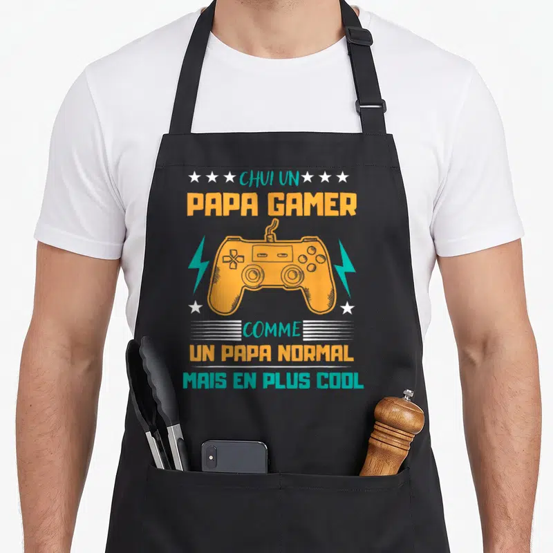 Tablier de cuisine noir avec illustration de manette de jeu vidéo orange et texte humoristique sur fond uni.