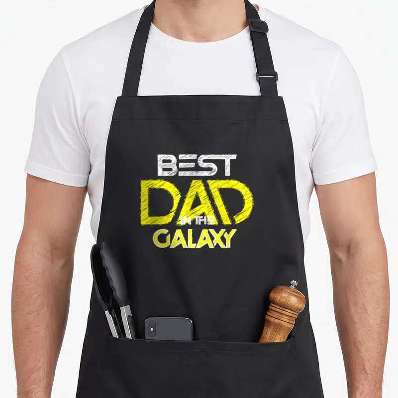 Tablier de cuisine noir avec inscription Best Dad in the Galaxy en lettres jaunes et blanches stylisées.