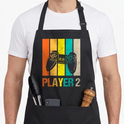Tablier de cuisine noir avec une manette de jeu vidéo sur fond de bandes colorées verticales orange jaune et turquoise.
