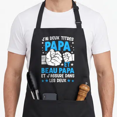 Tablier de cuisine noir avec l'inscription J'ai deux titres Papa et Beau-papa et une illustration de poignées de main.