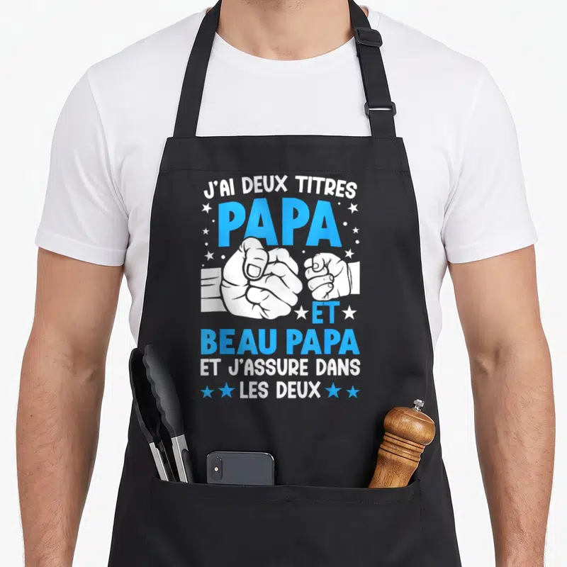 Tablier de cuisine noir avec l'inscription J'ai deux titres Papa et Beau-papa et une illustration de poignées de main.