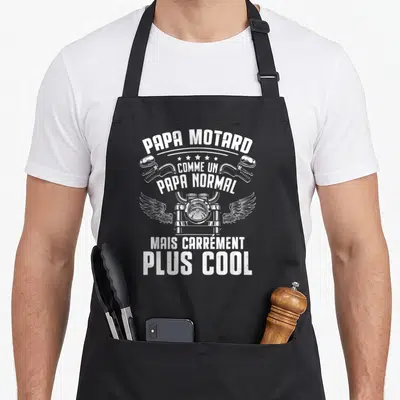Tablier de cuisine noir avec illustration blanche d'un phare de moto ailé et texte humoristique sur fond uni.