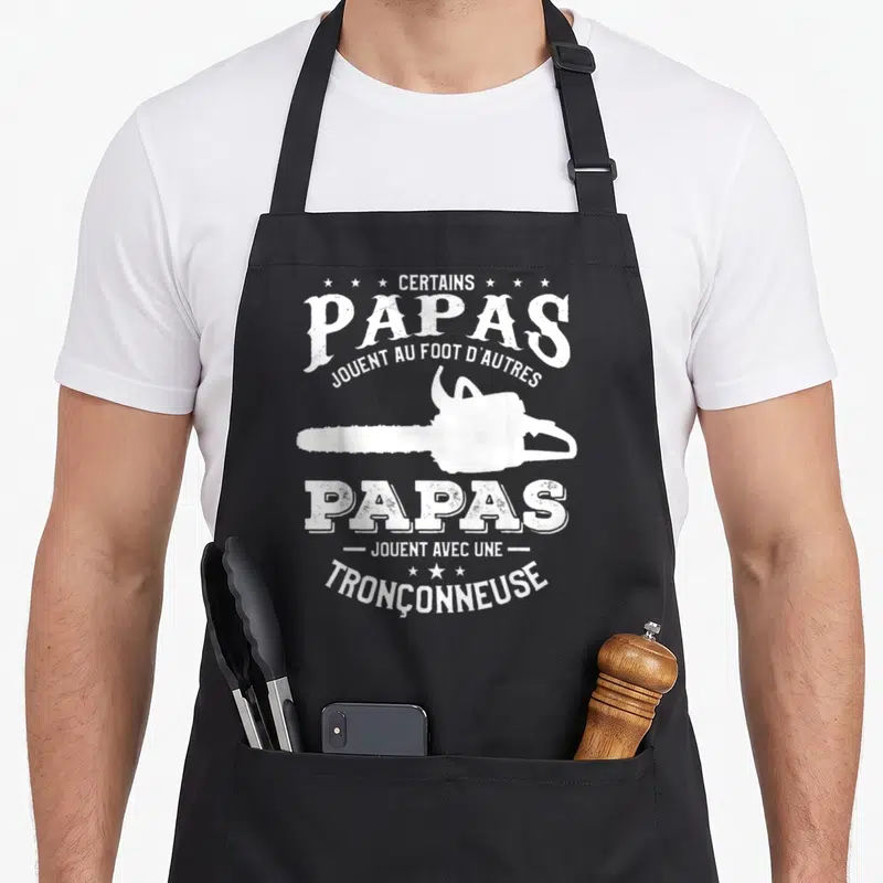 Tablier de cuisine noir avec silhouette blanche de tronçonneuse et texte certain papas jouent avec une tronçonneuse