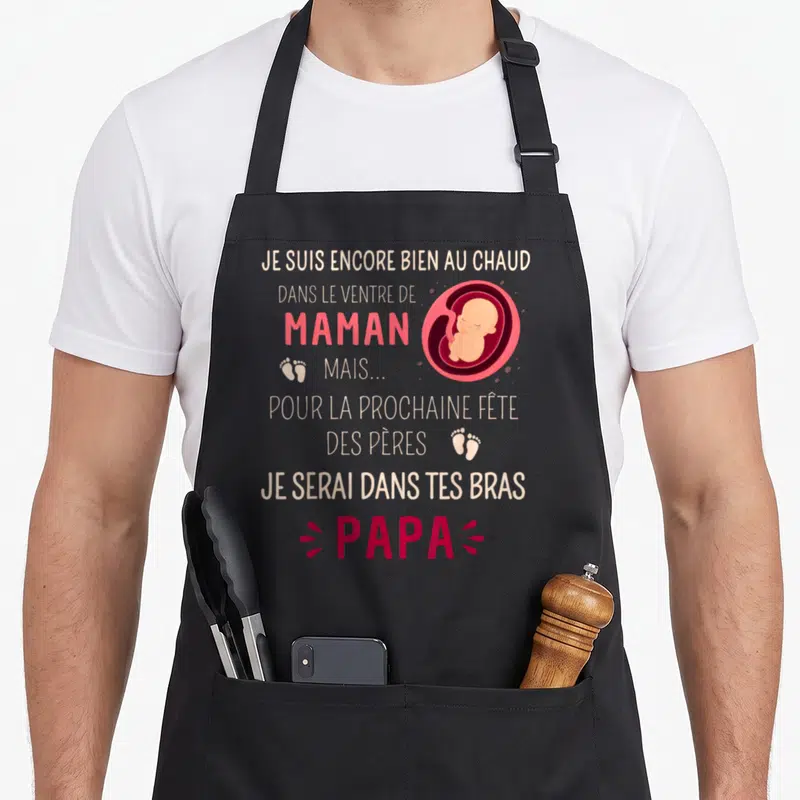 Tablier de cuisine noir avec texte humoristique sur la future fête des pères et illustration d'un bébé dans un cercle rose.