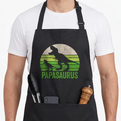 Tablier de cuisine noir avec illustration vintage Papasaurus montrant un grand et un petit dinosaure gris devant un soleil vert strié.