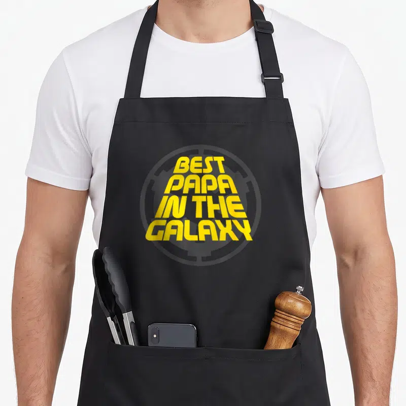 Tablier de cuisine noir avec logo circulaire gris et texte jaune Best Papa in the Galaxy au centre.