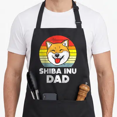 Tablier de cuisine noir avec un Shiba Inu souriant et linscription Shiba Inu Dad en blanc sur fond de soleil coloré.