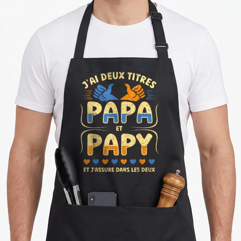 Tablier de cuisine noir avec l'inscription J'ai deux titres Papa et Papy et j'assure dans les deux accompagnée de mains et de cœurs.