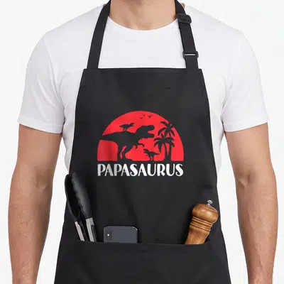 Tablier de cuisine noir avec silhouette T Rex et bébé dinosaure devant un soleil rouge et le mot Papasaurus.