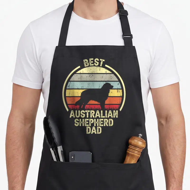 Tablier de cuisine noir avec silhouette Berger australien et texte vintage coloré sur fond de soleil couchant.