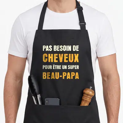 Tablier de cuisine noir avec l'inscription humoristique Pas besoin de cheveux pour être un super beau-père en lettres orange et crème.